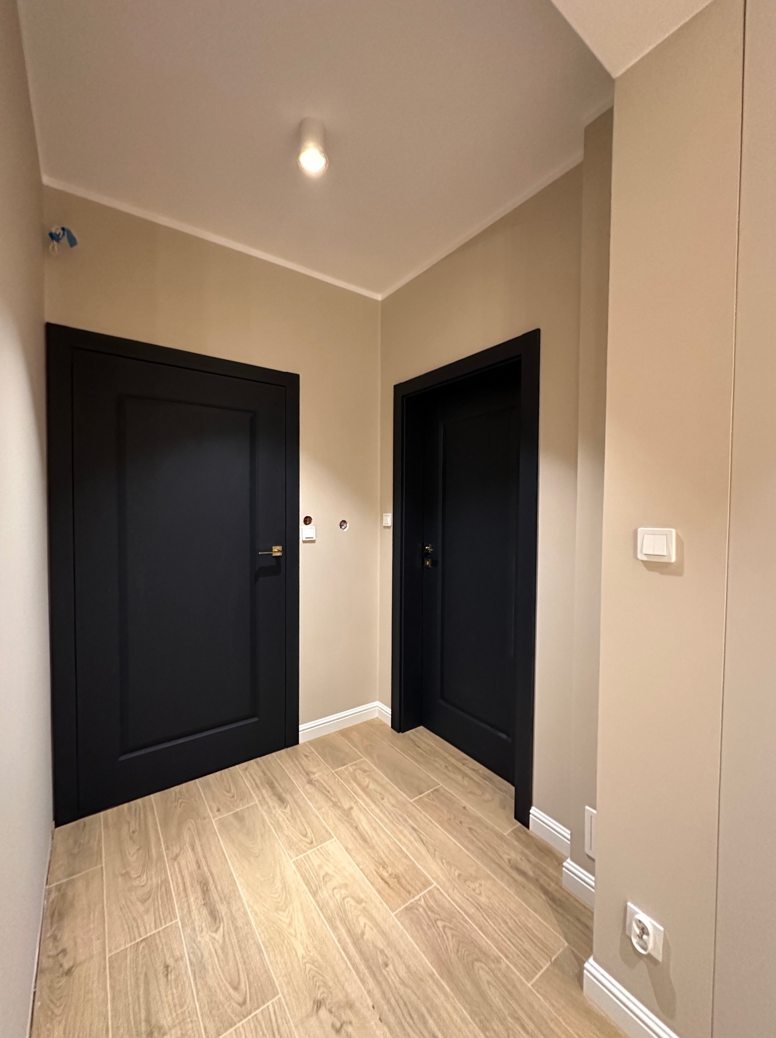 Korytarz po remoncie z dwiema czarnymi, nowoczesnymi drzwiami z złotymi klamkami, beżowe ściany, podłoga wyłożona panelami imitującymi drewno, białe listwy przypodłogowe i włączniki światła...
