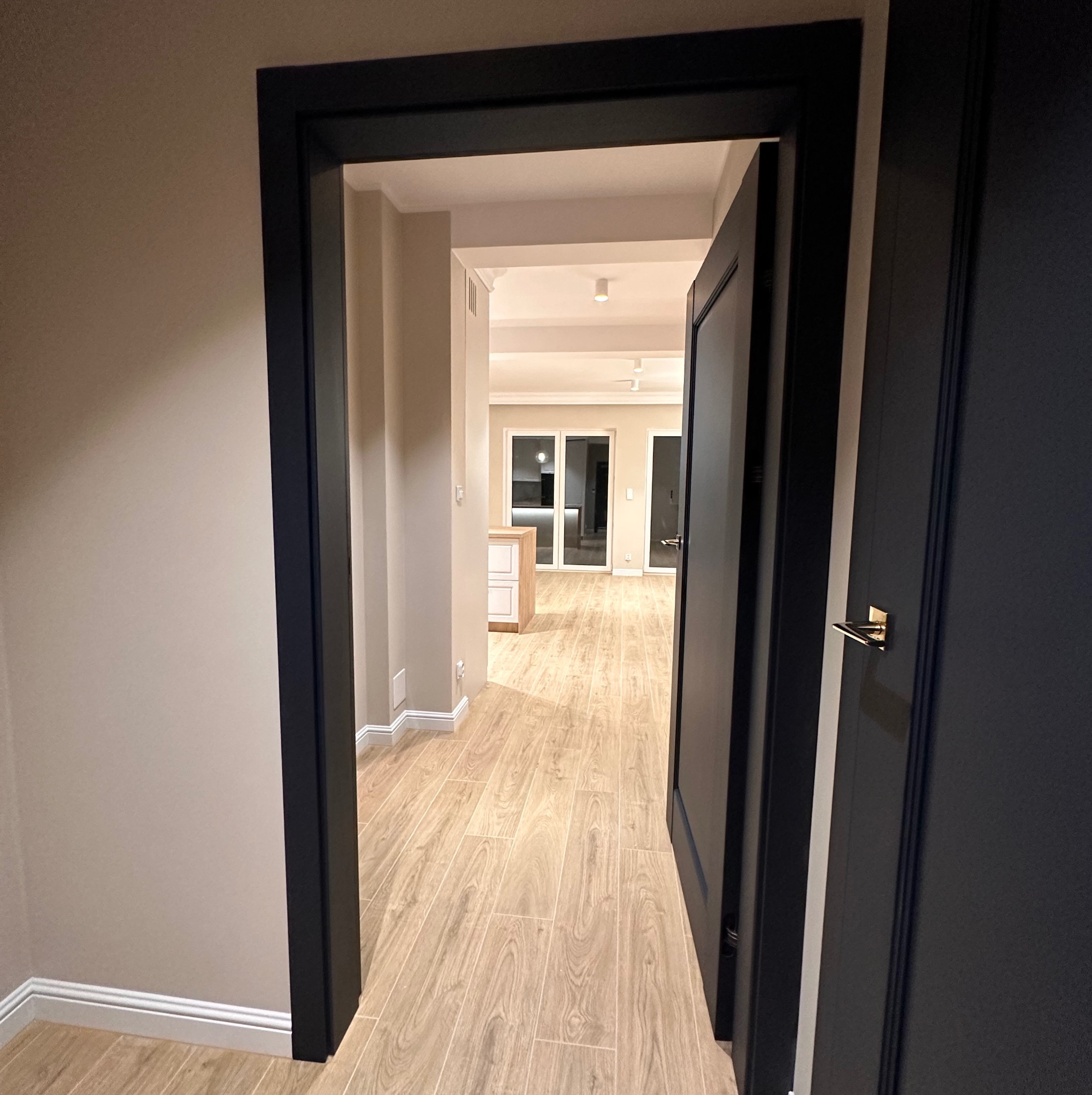 Widok z korytarza przez otwarte, czarne drzwi na wykończone wnętrze salonu z jasną podłogą imitującą drewno i białymi ścianami, w tle widoczne przeszklone drzwi balkonowe.