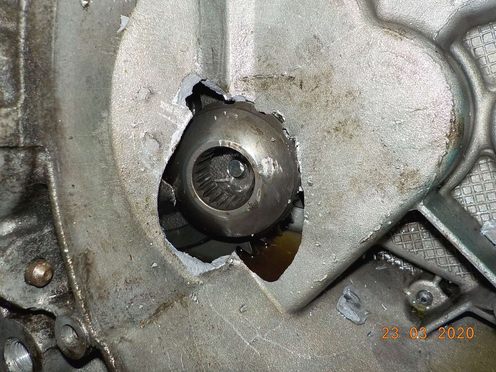 Uszkodzona obudowa skrzyni biegów z widocznym wyłamanym otworem i fragmentami metalu, wyciekający olej, data na zdjęciu 23.03.2020.