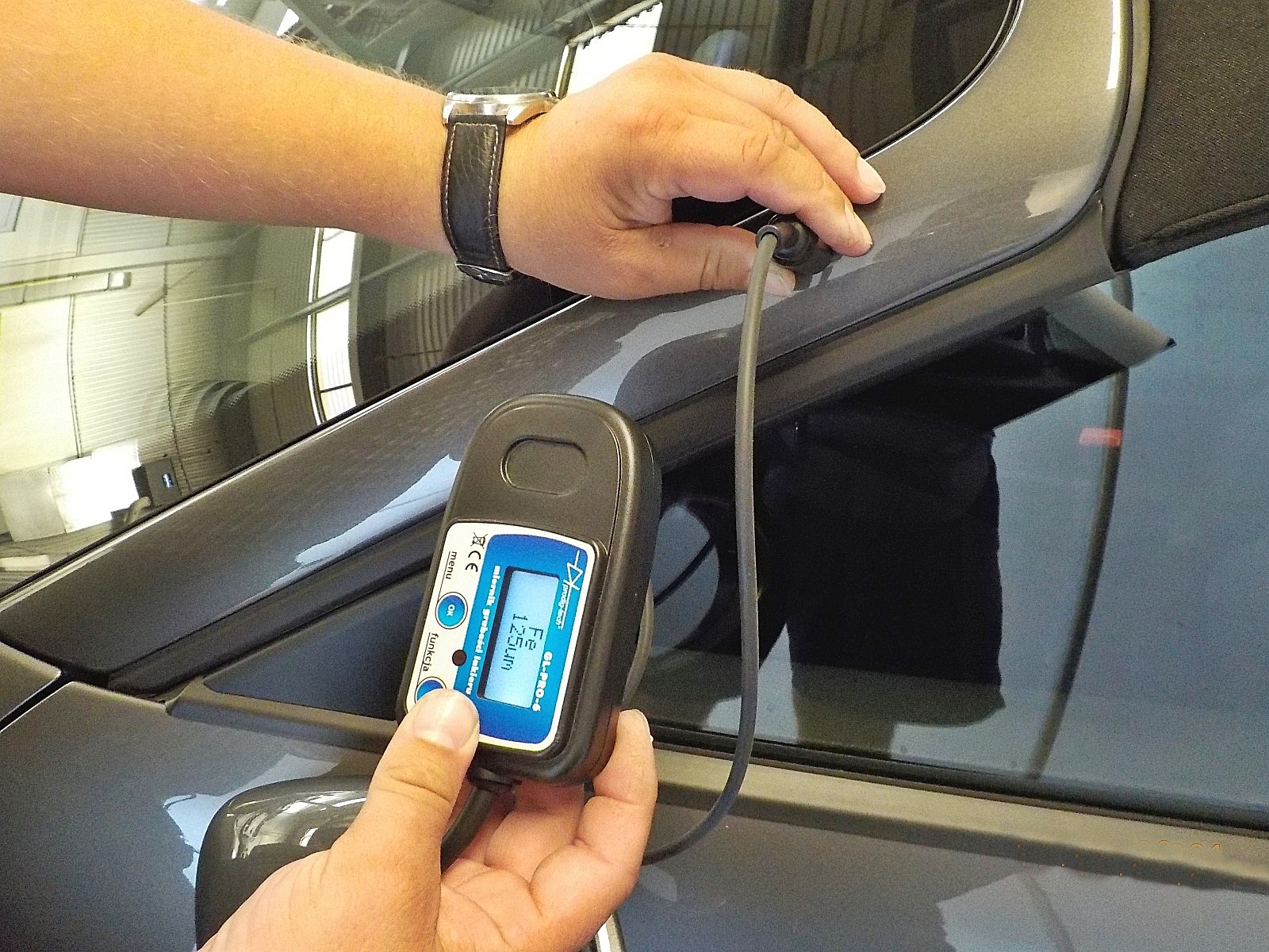 Miernik grubości lakieru przykładany do szarego samochodu z otwartym dachem, widoczny wyświetlacz urządzenia wskazujący wartość pomiaru.