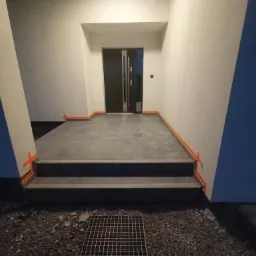 Układanie ciemnoszarych płytek na schodach wejściowych, zabezpieczonych pomarańczową taśmą, widoczna krata ściekowa i drobny żwir.