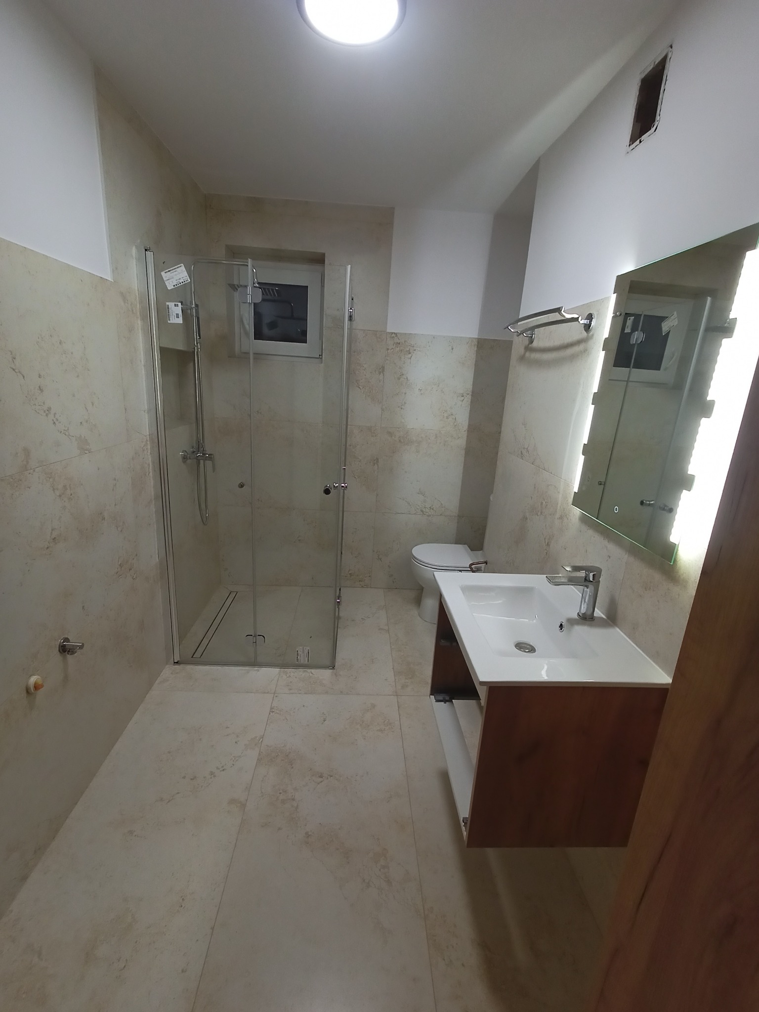 Wykończona łazienka z kabiną prysznicową ze szklanymi drzwiami, umywalką na szafce z drewnianym frontem i podświetlanym lustrem, wykończona płytkami w odcieniach beżu.