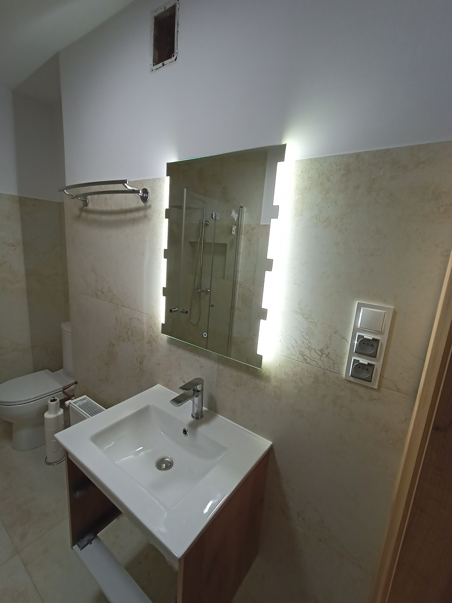 Nowoczesna łazienka z podświetlanym lustrem, umywalką nablatową, toaletą i grzejnikiem, wykończona płytkami imitującymi marmur.