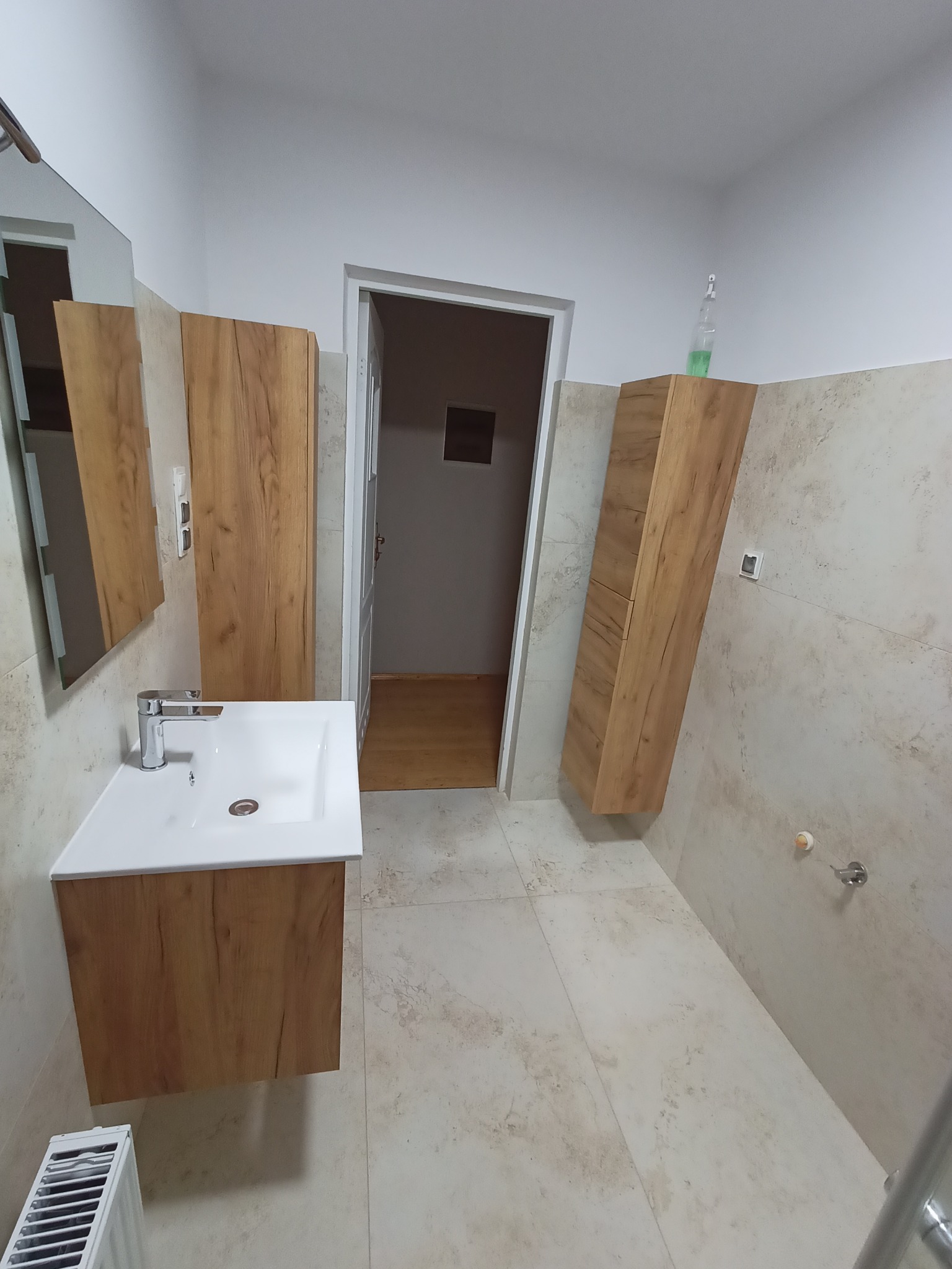 Nowoczesna łazienka z beżowymi płytkami, wiszącą szafką pod umywalką z drewnianym wykończeniem, lustrem i wysoką szafką wiszącą po prawej stronie, widok na drzwi do innego pomieszczenia.