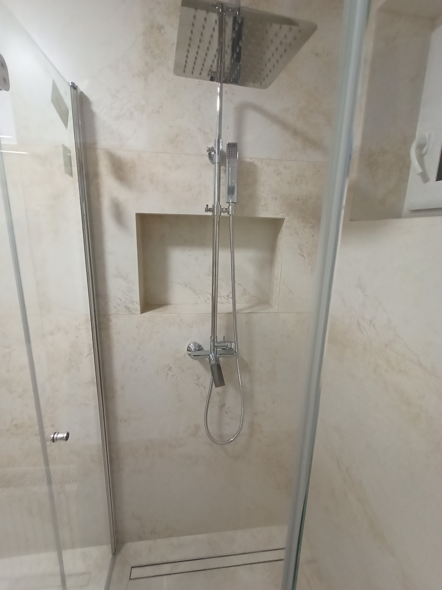 Nowoczesna kabina prysznicowa z deszczownicą i wnęką w ścianie wykończona beżowymi płytkami imitującymi marmur, widoczny odpływ liniowy.