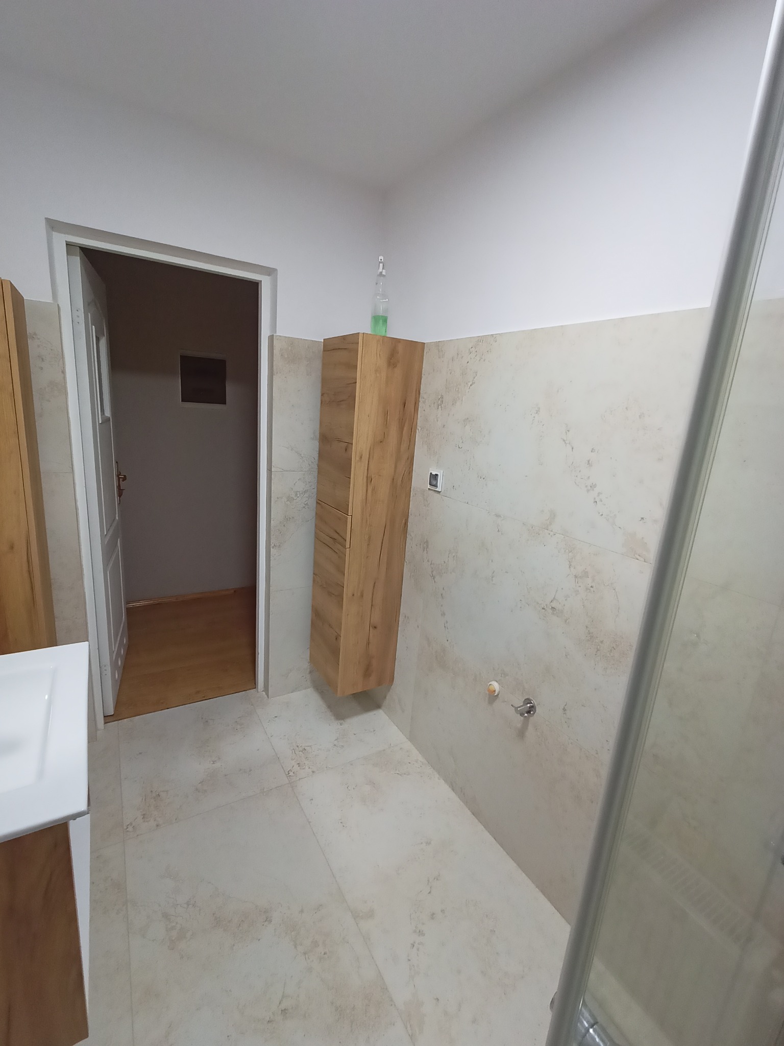 Jasna łazienka z beżowymi płytkami imitującymi marmur na ścianach i podłodze. Widoczna wisząca szafka z drewnopodobnym dekorem, częściowo widoczna kabina prysznicowa z metalową ramą oraz fragment...