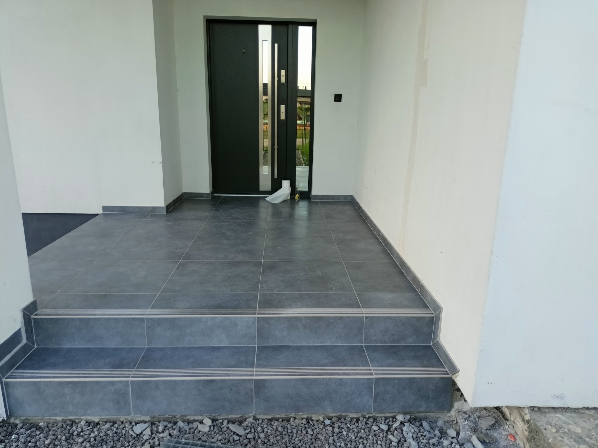 Wykończone płytki ceramiczne na schodach wejściowych do domu, z widoczną nowoczesną, grafitową stolarką drzwiową i fragmentem elewacji w kolorze złamanej bieli. Widoczne fugi i listwy...