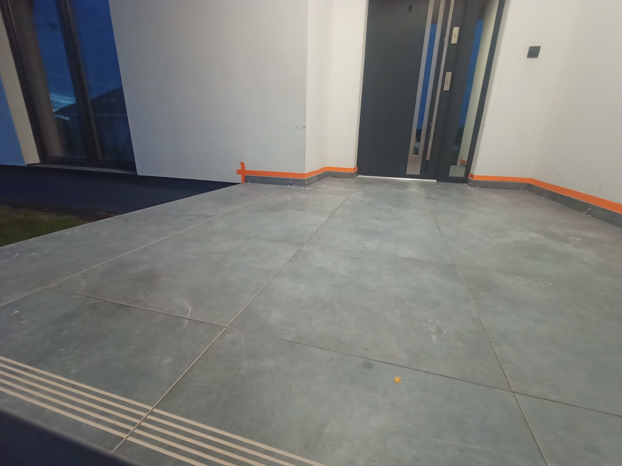 Zewnętrzny taras wyłożony szarymi, kwadratowymi płytkami z pasami antypoślizgowymi na krawędzi, zabezpieczony pomarańczową taśmą malarską przy ścianie i drzwiach wejściowych.