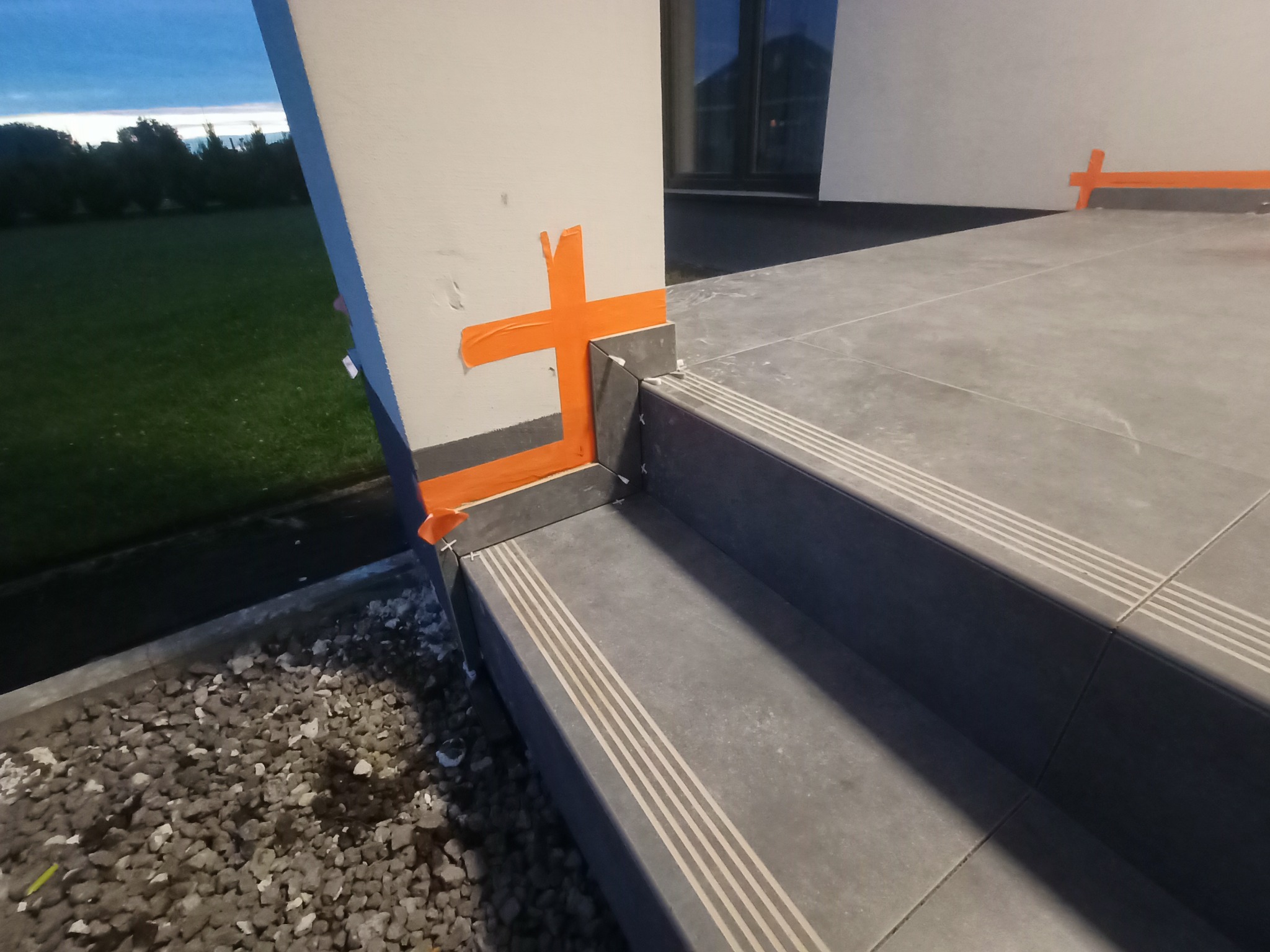 Montaż płytek na schodach zewnętrznych, widoczne pomarańczowe taśmy maskujące na ścianie i płytkach, szare płytki z antypoślizgowymi paskami, otoczenie z kamyków.