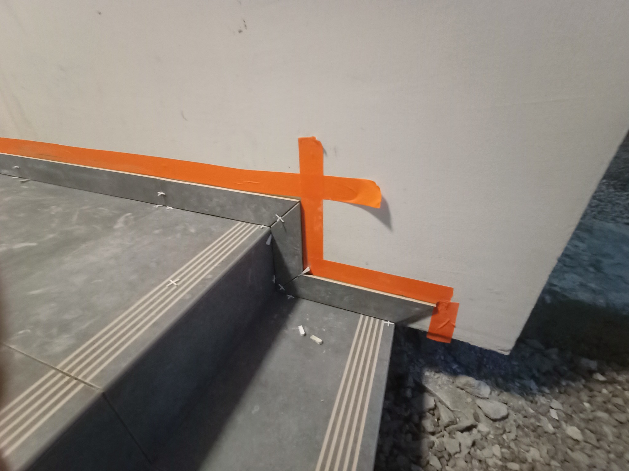 Szare płytki schodowe z czterema jasnymi paskami, zabezpieczone pomarańczową taśmą malarską przy ścianie, widoczne krzyżyki dystansowe i gruz na podłodze.