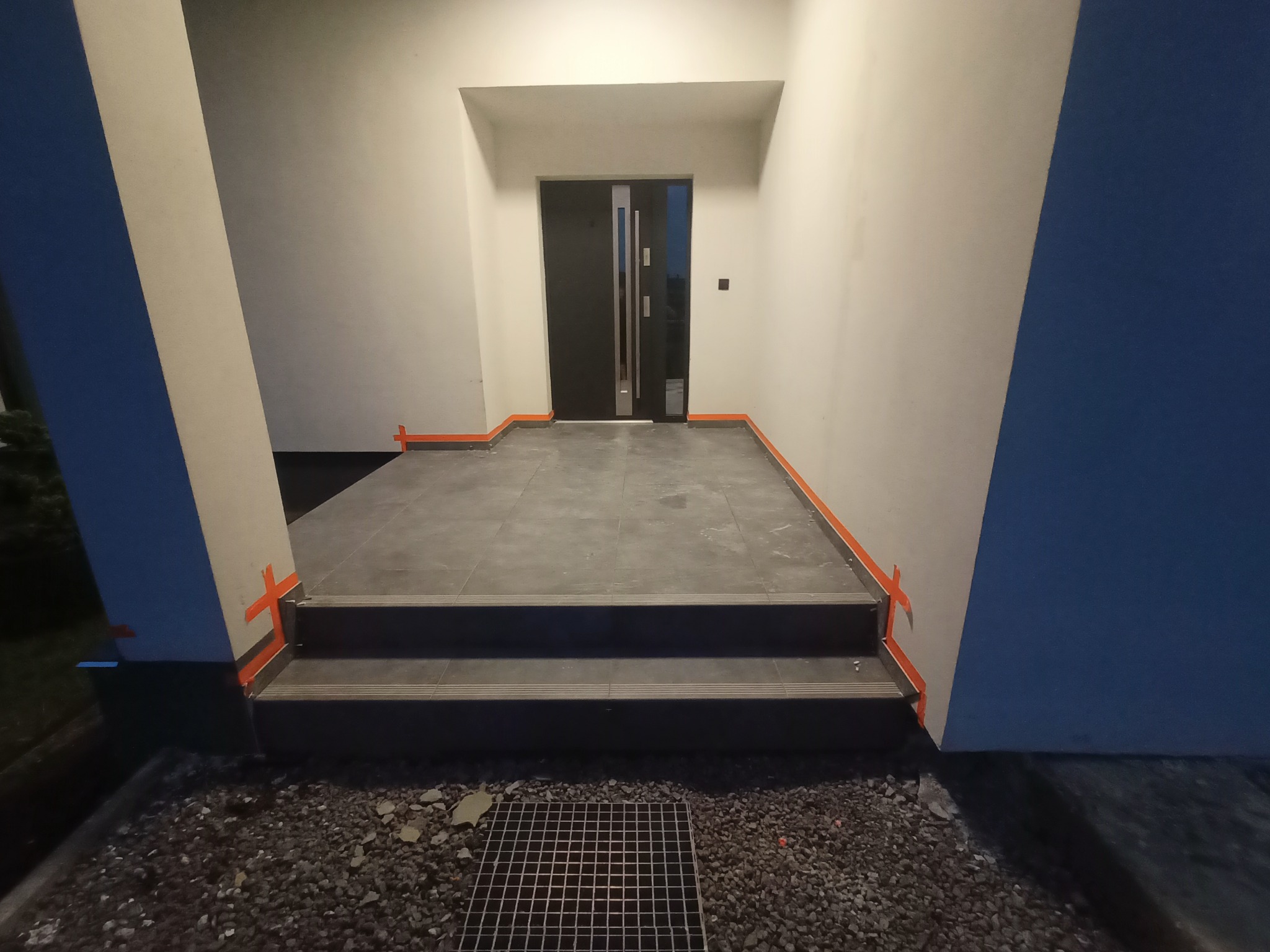 Układanie ciemnoszarych płytek na schodach wejściowych, zabezpieczonych pomarańczową taśmą, widoczna krata ściekowa i drobny żwir.