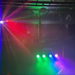 Scena oświetlona kolorowymi reflektorami i laserami, z drewnianą podłogą i sprzętem nagłośnieniowym w tle