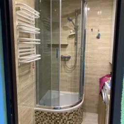 Narożna kabina prysznicowa z mozaiką u dołu, zamontowana w łazience wykończonej beżowymi płytkami, widoczny grzejnik drabinkowy i pralka w tle.