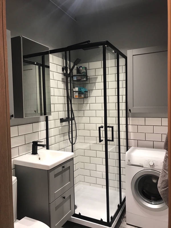 Nowoczesna łazienka z kabiną prysznicową w czarnej ramie, umywalką z szafką w odcieniach szarości i pralką. Ściany wyłożone białymi, prostokątnymi płytkami imitującymi cegłę.
