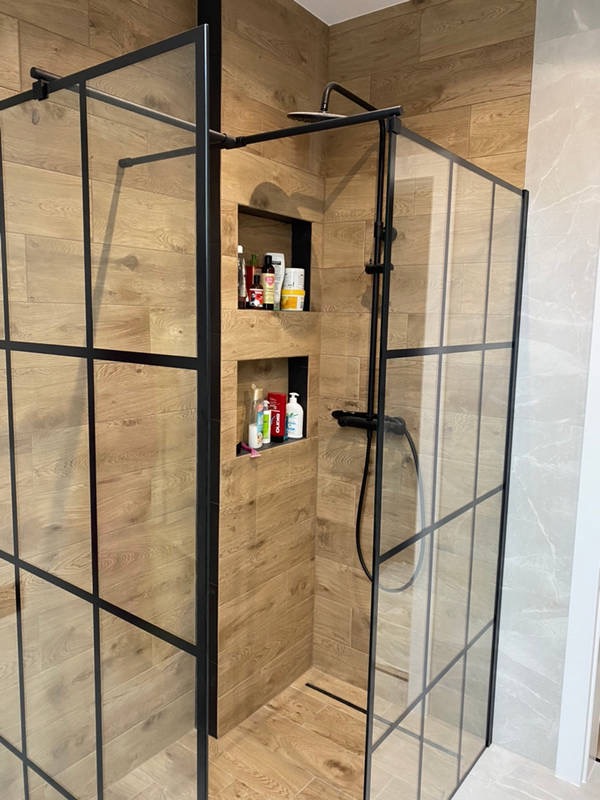 Nowoczesna kabina prysznicowa typu walk-in z czarnymi profilami i drewnopodobnymi płytkami, wbudowane półki z kosmetykami, odpływ liniowy w podłodze.