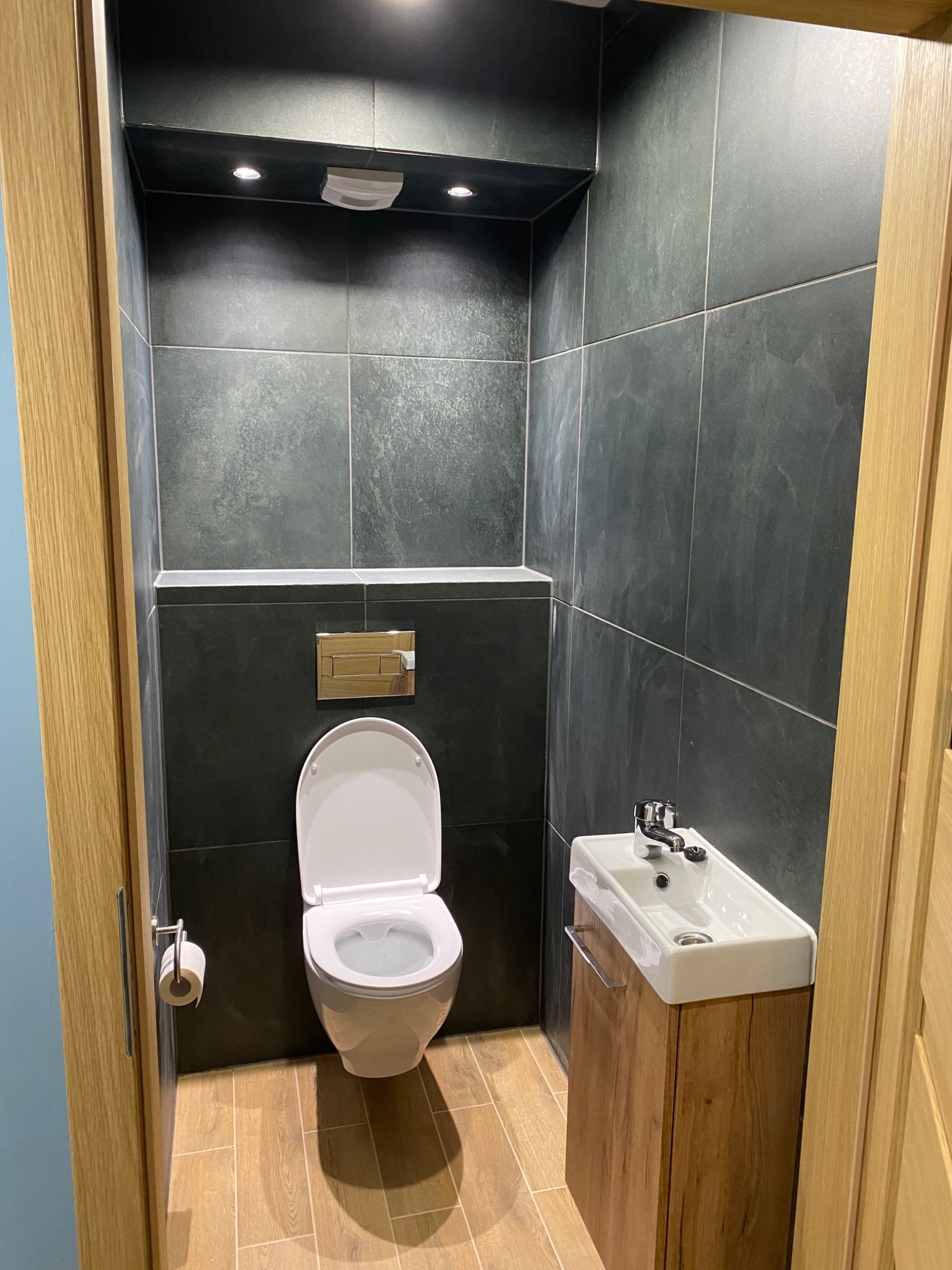 Nowoczesna łazienka z ciemnymi płytkami imitującymi kamień, podwieszana toaleta z chromowanym przyciskiem spłukiwania, umywalka nablatowa na szafce z drewnopodobnym fornirem i drewniane obramowanie...