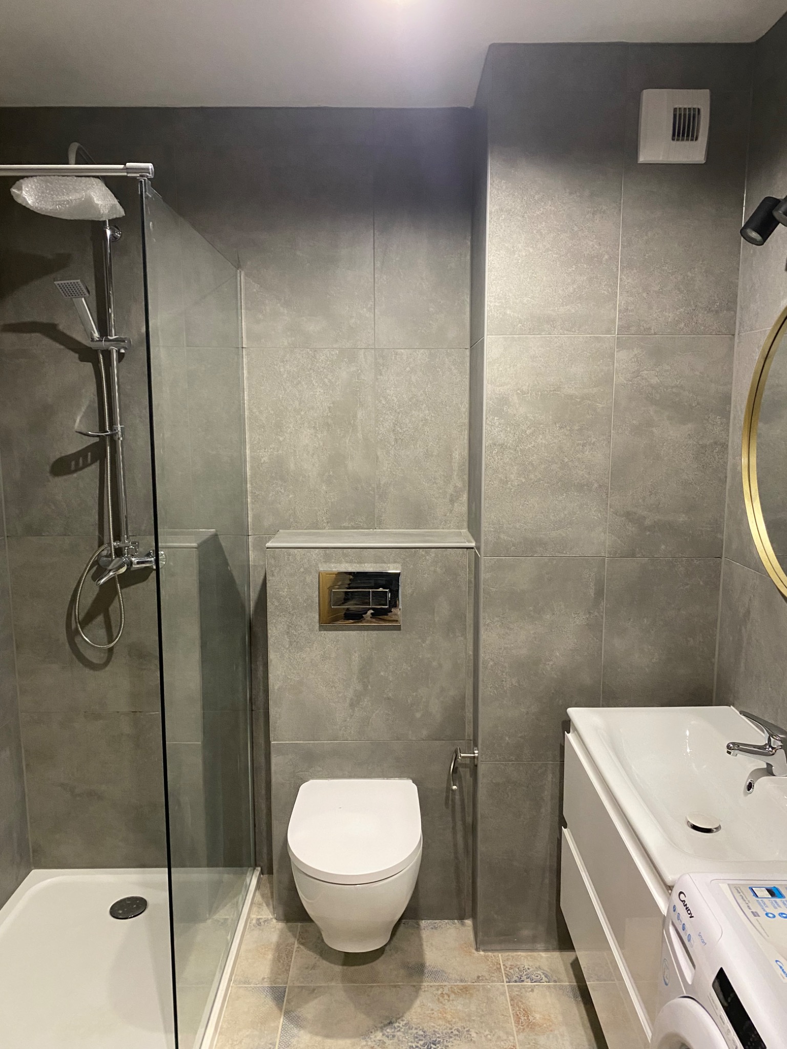 Nowoczesna łazienka z szarymi płytkami, kabiną prysznicową z osłoną z folii, wiszącą toaletą, umywalką z szafką i pralką Candy.