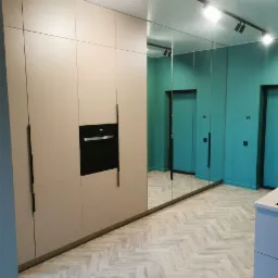 Szafa wnękowa w odcieniach beżu z wbudowanym piekarnikiem, przylegająca do ściany wyłożonej lustrami, odbijająca turkusowe ściany i parkiet ułożony w jodełkę.