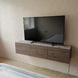 Telewizor zawieszony na ścianie nad szafką RTV z drewnianymi frontami i szarym blatem, widoczne kable, ściana w beżowym odcieniu z delikatnym wzorem.