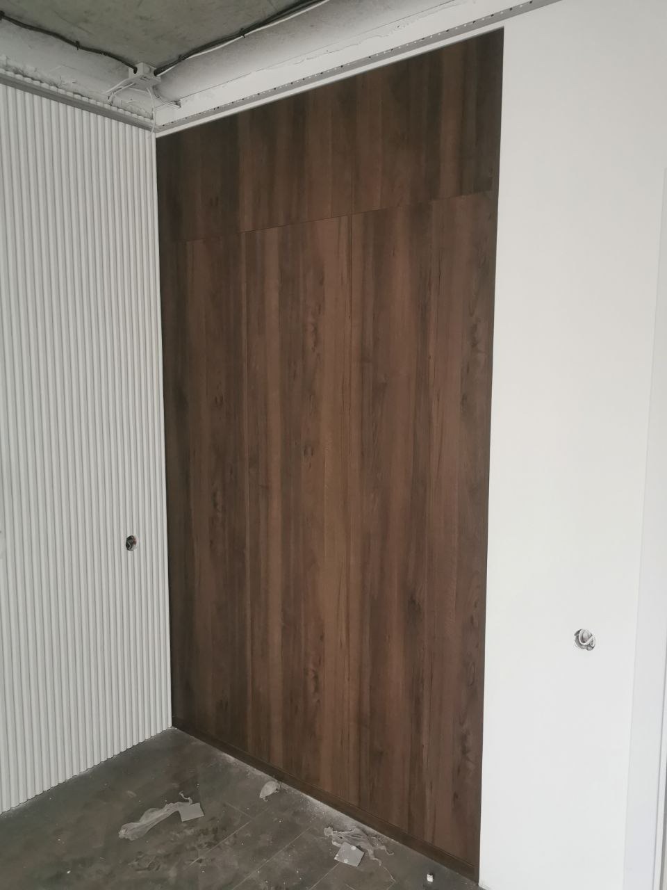 Wnętrze w trakcie wykańczania: brązowa zabudowa meblowa z panelami imitującymi drewno, połączona z białą ścianą i dekoracyjną ścianą z pionowymi żłobieniami, widoczne niedokończone podłogi...