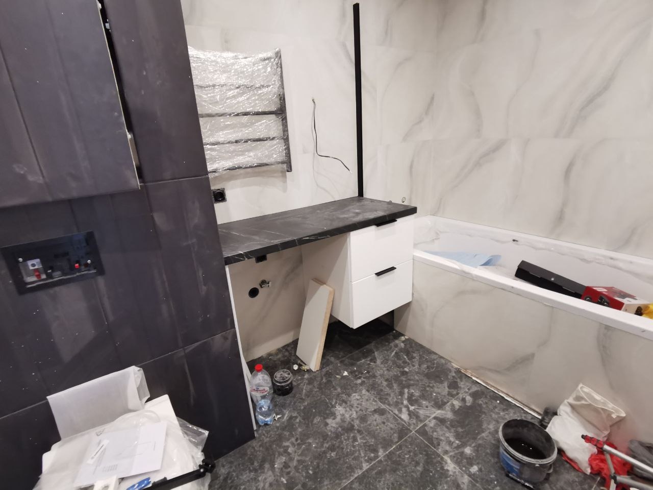 Niedokończona łazienka z czarnym blatem, białą szafką z dwiema szufladami, wanną w marmurowej obudowie i grzejnikiem drabinkowym owiniętym folią. Widoczne ślady prac remontowych, w tym butelka wody...