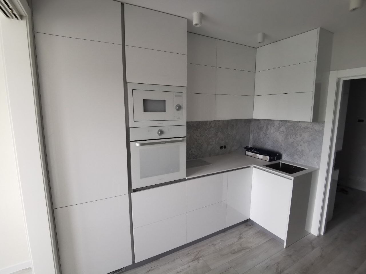 Nowoczesna, minimalistyczna kuchnia z białymi, lakierowanymi frontami szafek, wbudowanym piekarnikiem i mikrofalówką, szarym blatem i panelami ściennymi imitującymi beton, oraz drewnianą podłogą.