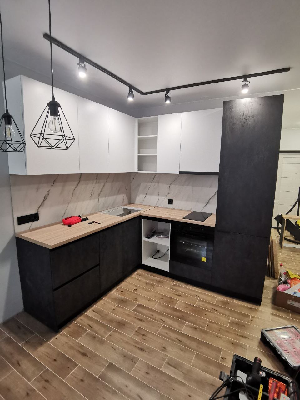 Nowoczesna kuchnia z czarnymi matowymi szafkami dolnymi i białymi górnymi, drewniany blat, marmurowe backsplash, oświetlenie szynowe i geometryczne lampy wiszące, widoczne narzędzia i elementy...