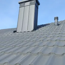 Szary dach z blachy trapezowej z kominem wentylacyjnym i odpowietrznikiem w słoneczny dzień.