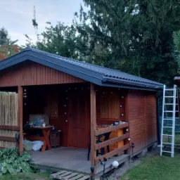 Drewniana altana z ciemnym dachem i elementami dekoracyjnymi, częściowo osłonięta bambusową przesłoną, obok drabina oparta o mały drewniany budynek, otoczona zielenią.