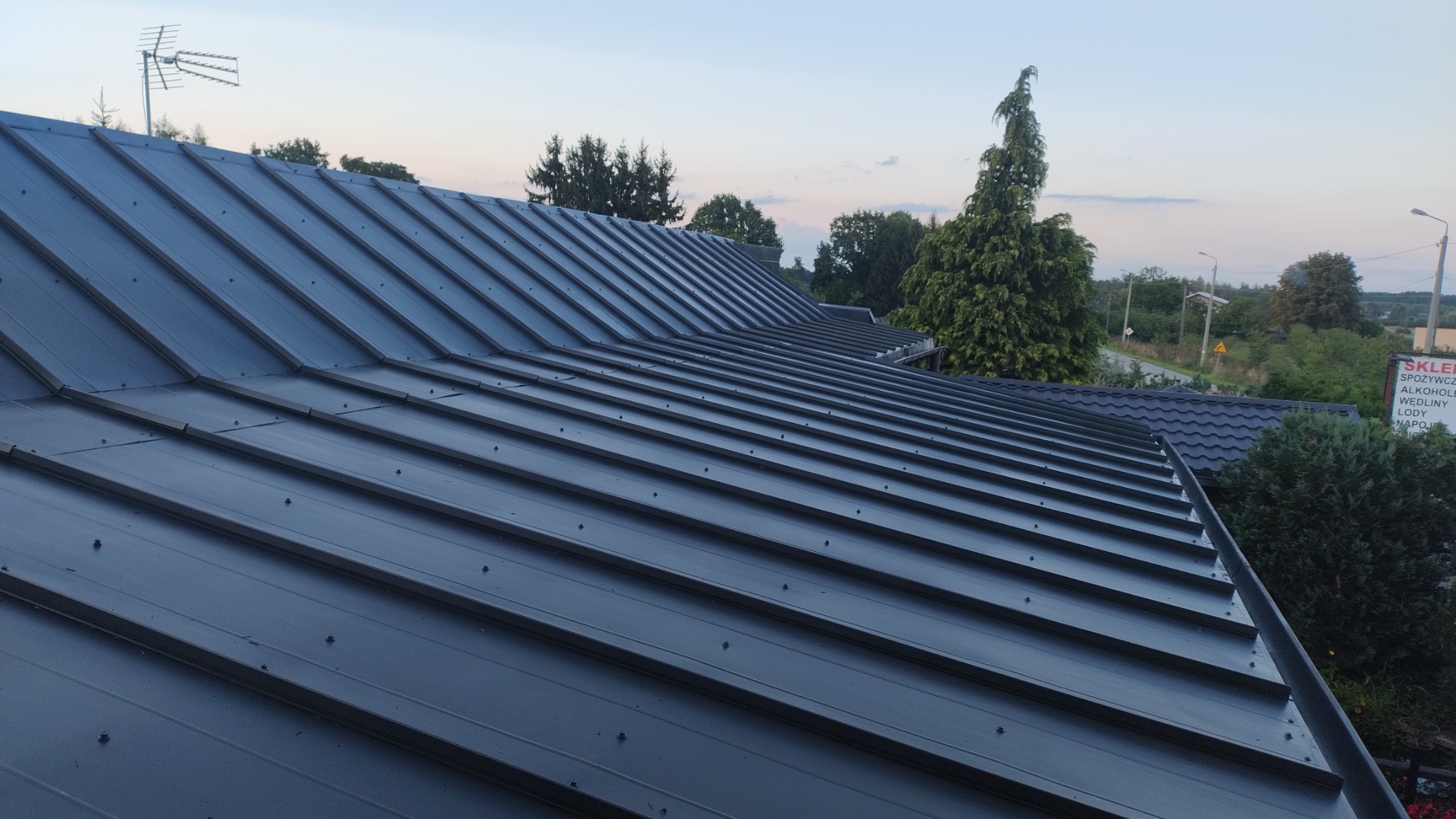 Nowy, ciemny blaszany dach z widocznymi mocowaniami, antena na szczycie, widok na okolicę z zielenią i budynkami w tle.
