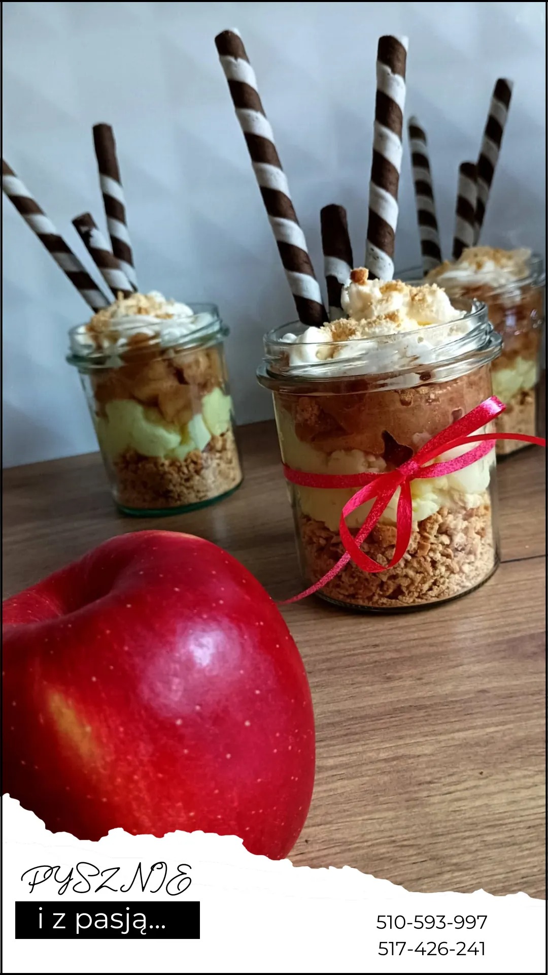 Trzy desery warstwowe w słoiczkach, udekorowane bitą śmietaną, posypką i rurkami waflowymi, jeden ze słoiczków przewiązany czerwoną wstążką, obok leży czerwone jabłko.