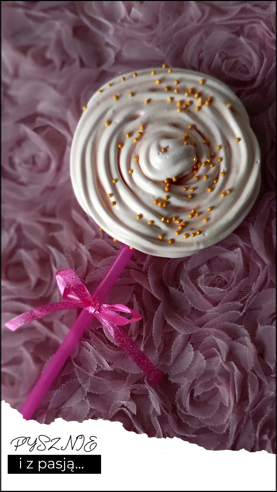 Biały lizak bezowy z posypką zlotych kuleczek na różowym tle z tkaniny w róże, różowa słomka z kokardką