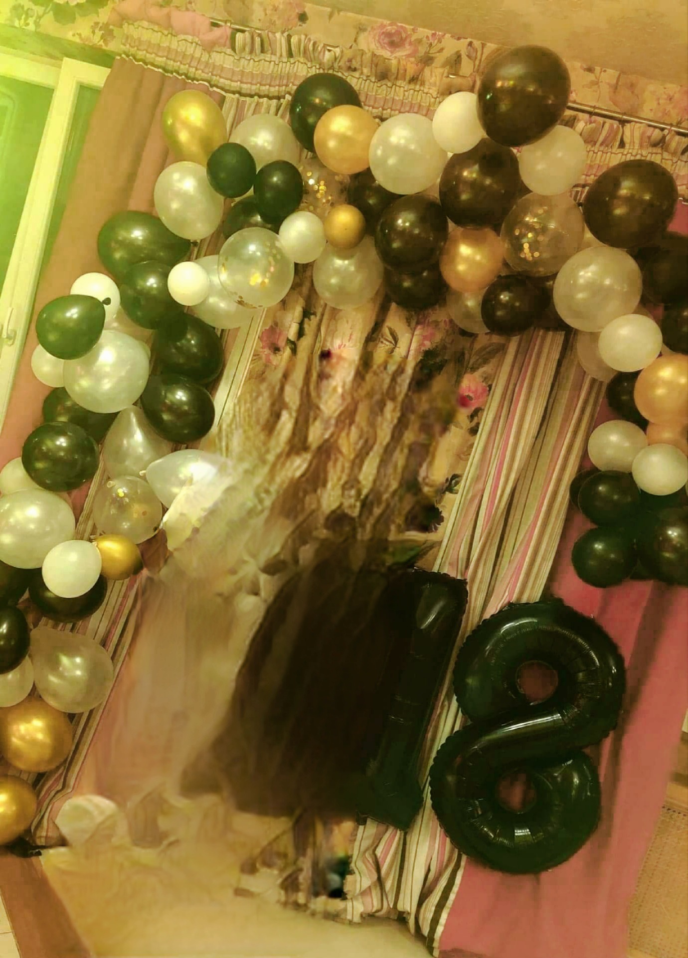 Aranżacja z balonów w odcieniach bieli, złota i zieleni, tworząca łuk na tle wzorzystej tkaniny, z foliowymi balonami w kształcie cyfr '1' i '8'.