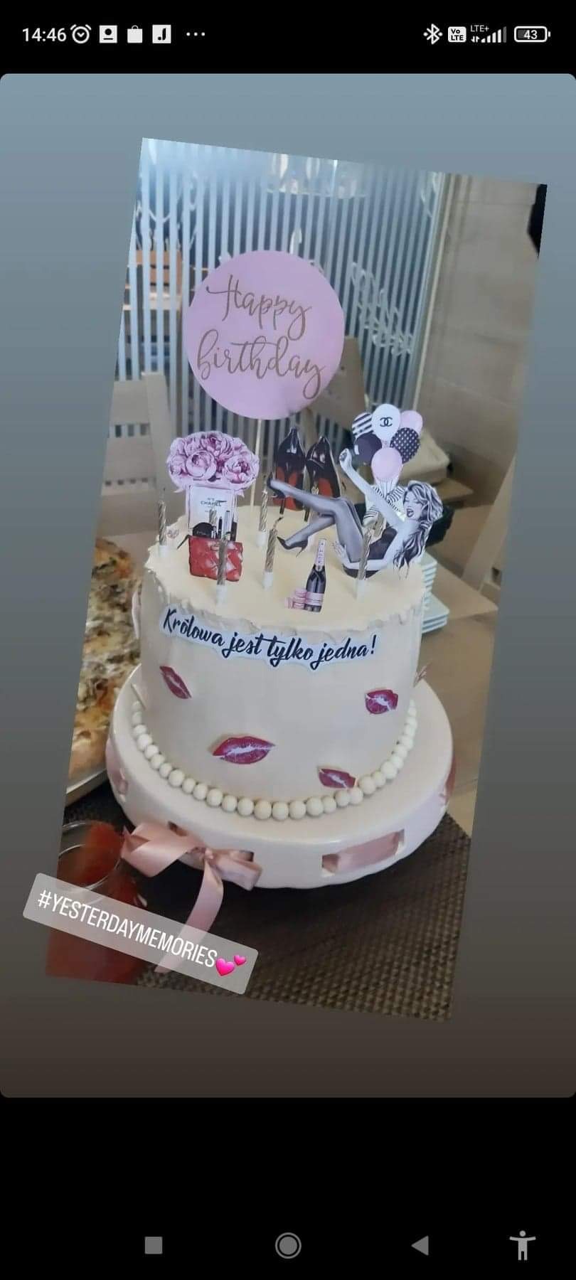 Dwupoziomowy tort urodzinowy w jasnych barwach, udekorowany napisami, motywami ust, butelek szampana, perfum, kwiatów i sylwetką kobiety, umieszczony na paterze z różową wstążką.