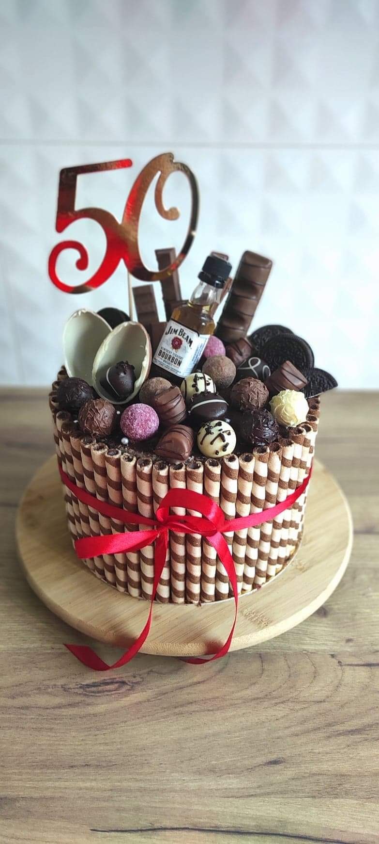 Tort urodzinowy udekorowany waflami, czekoladkami, mini butelką bourbona Jim Beam i topperem z liczbą 50, na drewnianej tacy.