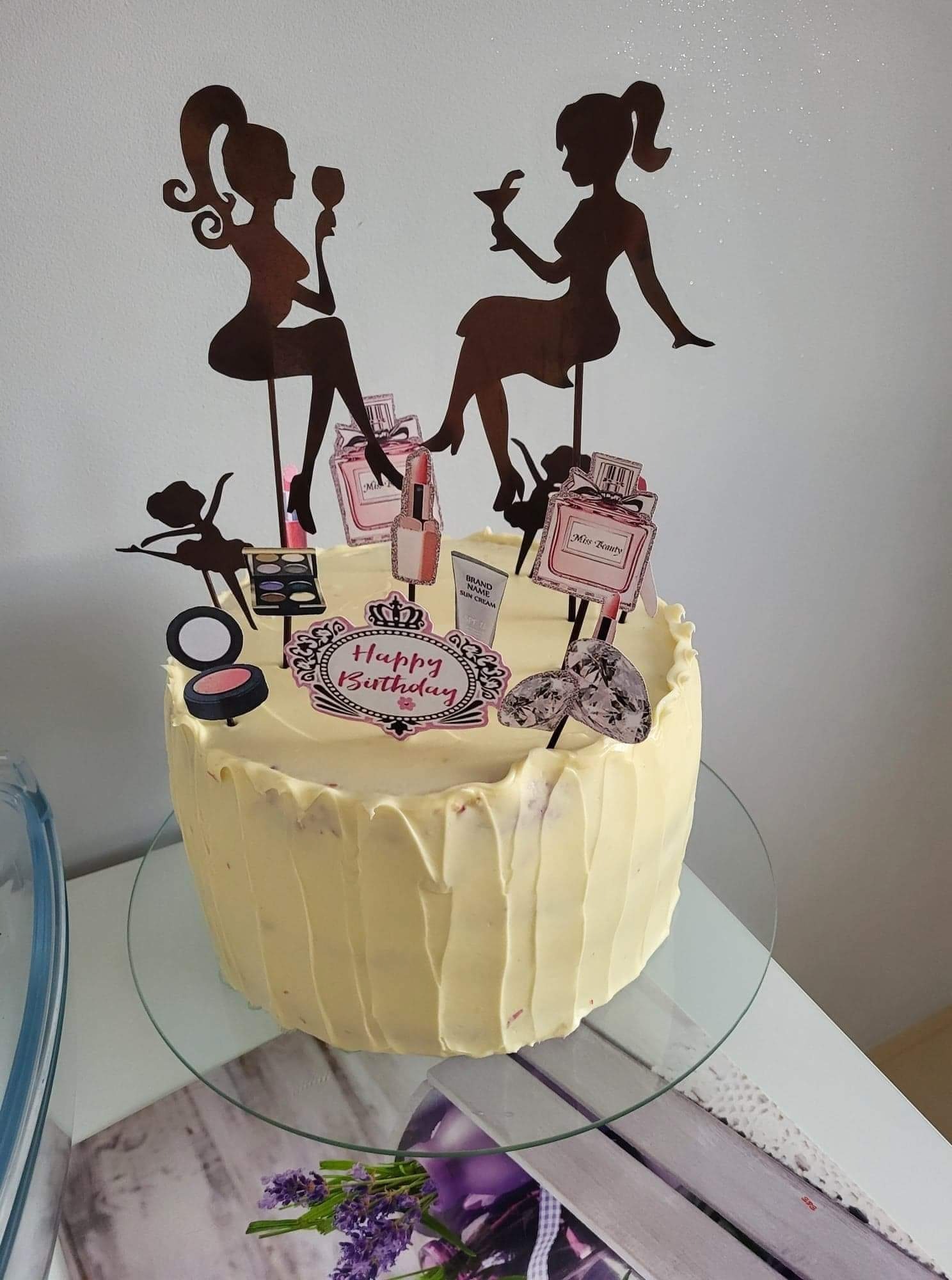 Tort urodzinowy z jasnym kremem, udekorowany topperami przedstawiającymi sylwetki kobiet z drinkami, kosmetyki i baletnicę, umieszczony na szklanej paterze.