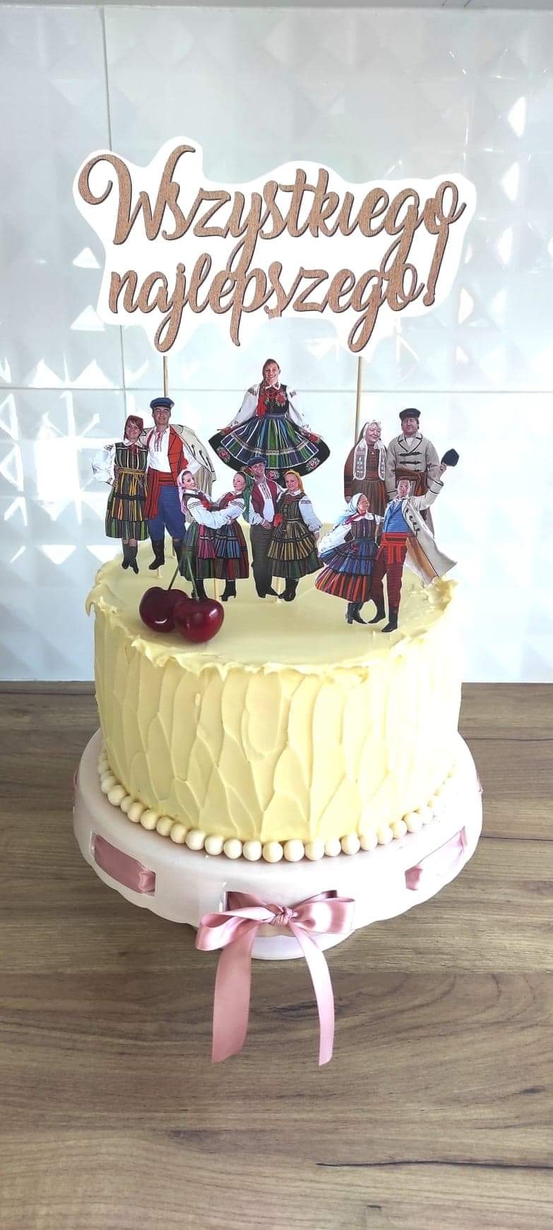 Tort z kremem w odcieniu wanilii udekorowany wiśniami i papierowymi postaciami w strojach ludowych, zwieńczony napisem 'Wszystkiego najlepszego!'