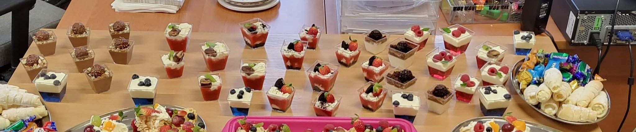 Szeroki kadr stołu z różnorodnymi deserami w małych, przezroczystych pojemnikach i na srebrnych paterach, w tle widoczny komputer stacjonarny i sterta serwetek.