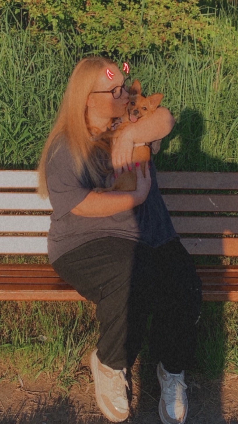 Kobieta w okularach siedzi na ławce w parku, całując małego, brązowego psa w pysk, pies uśmiecha się, a na głowie kobiety widoczne są cyfrowe rogi diabła.