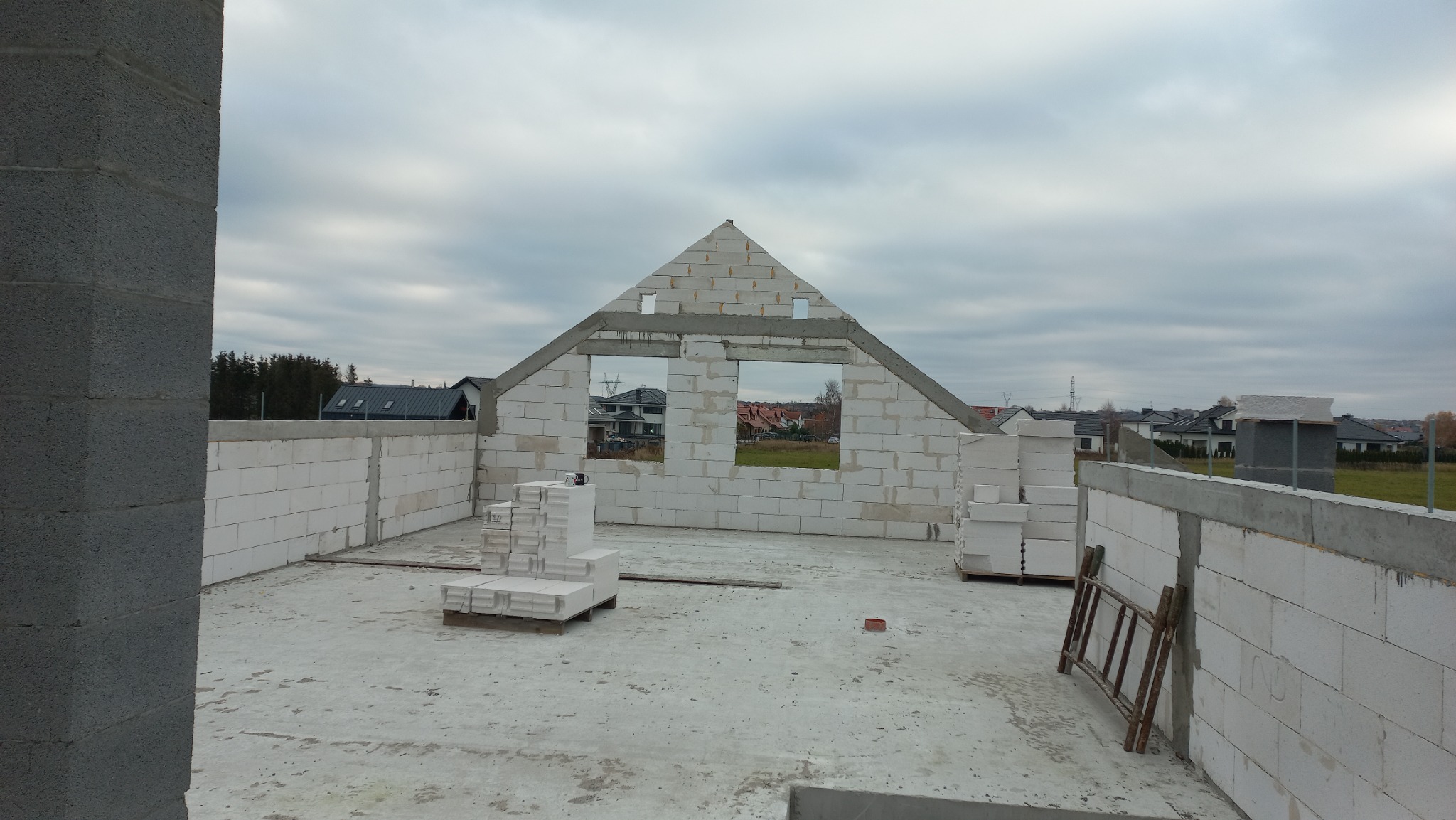 Wznoszenie ścian domu murowanego w Szczecinie z bloczków betonu komórkowego, widoczne puste otwory okienne i szczyt dachu, na podłodze palety z bloczkami i drabina oparta o ścianę.