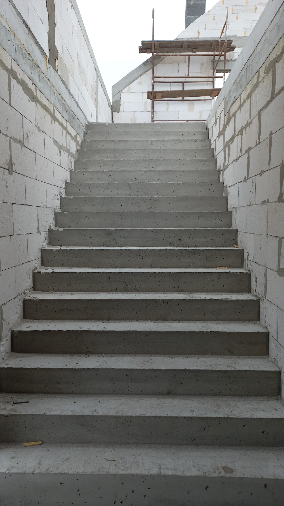 Surowe, betonowe schody prowadzące w górę, pomiędzy ścianami z białych bloczków, z widocznym rusztowaniem w tle, w trakcie budowy domu.