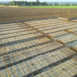 Zbrojenie stalowe ułożone na bloczkach betonowych w przygotowaniu do wylania fundamentów, widok z góry na tle zaoranego pola.