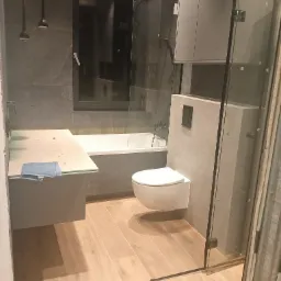 Nowoczesna łazienka w trakcie wykańczania: szare płytki ścienne, drewnopodobna podłoga, wisząca toaleta, szklana kabina prysznicowa, blat z niebieską ścierką zabezpieczony folią, minimalistyczne...