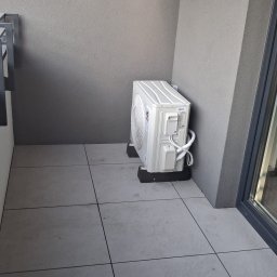 EMES Wykończenia wnętrz - Biały klimatyzator zewnętrzny na szarym balkonie, widok z boku. Minimalistyczny design, nowoczesne wykończenie, widoczne przewody.