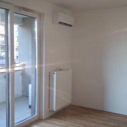 EMES Wykończenia wnętrz - Jasne wnętrze z klimatyzatorem na ścianie, parkietem i dużym oknem balkonowym. Widoczny grzejnik pod klimatyzatorem. Minimalistyczny design.
