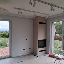 EMES Wykończenia wnętrz - Wnętrze w trakcie wykańczania: kominek obudowany beżową płytą, szyny z reflektorami na suficie, duże okna z widokiem na zieleń, podłoga zabezpieczona folią.