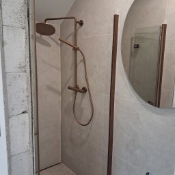 EMES Wykończenia wnętrz - Nowoczesna kabina prysznicowa z deszczownicą w odcieniach brązu, szklane drzwi z brązowymi ramami, okrągłe lustro i szare płytki. Częściowo widoczny surowy mur.