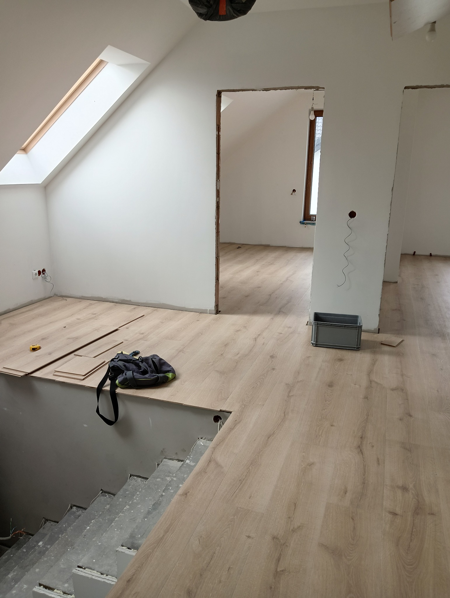 Poddasze w trakcie remontu, widoczne panele podłogowe, torba narzędziowa, poziomica, schody betonowe, surowe ściany i przygotowane otwory drzwiowe.