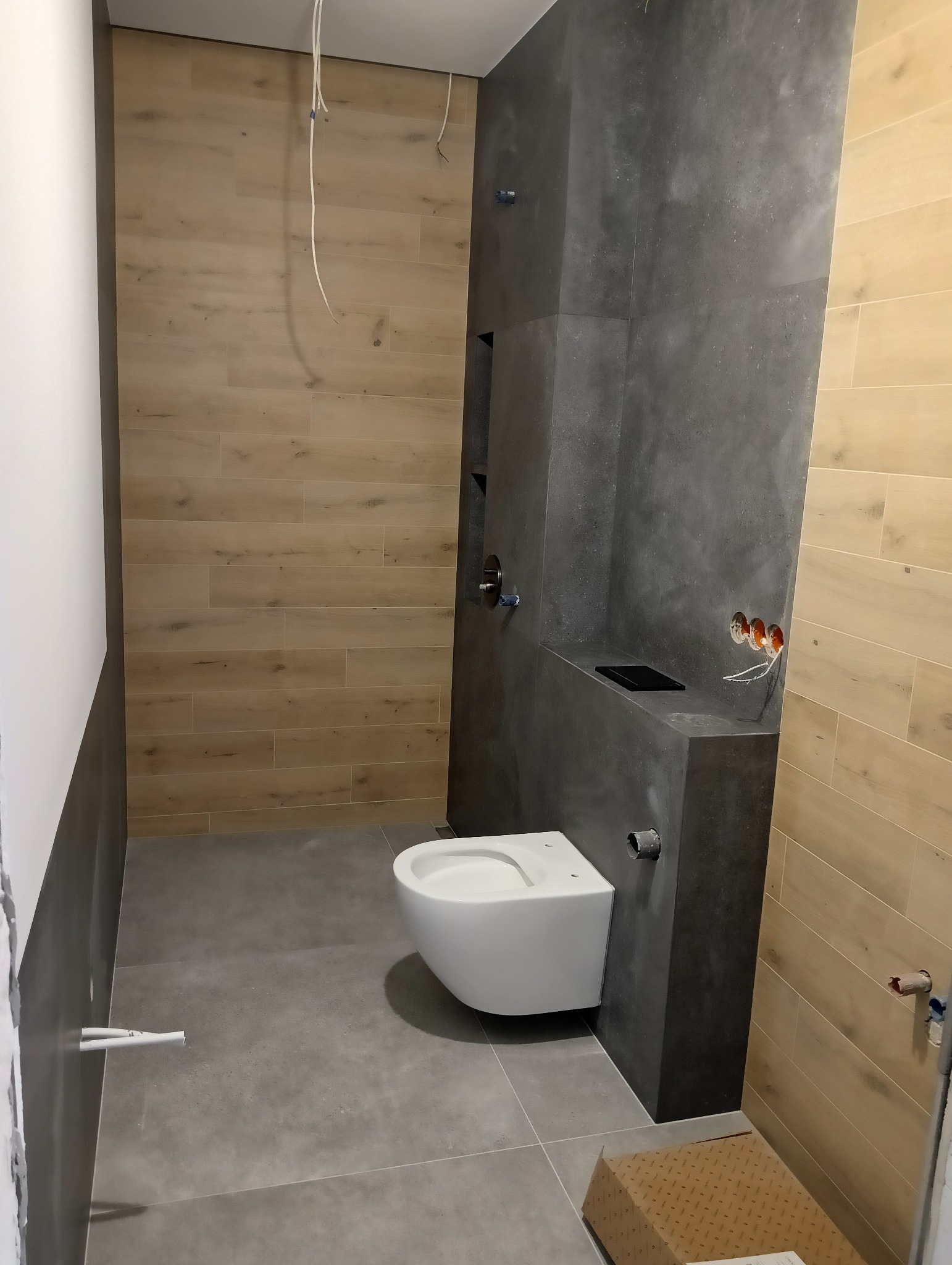 Wnętrze łazienki w trakcie wykańczania: ściany wyłożone płytkami imitującymi drewno i szarym betonem, wisząca miska WC, wystające kable elektryczne i rury hydrauliczne, karton na podłodze.