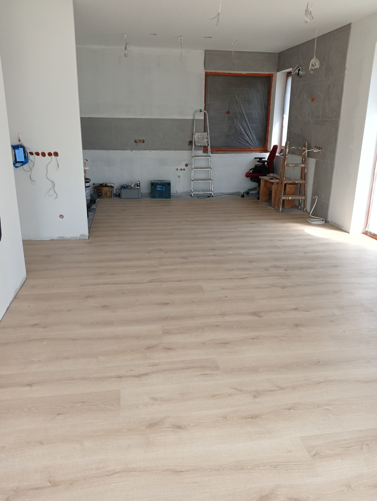 Jasne panele podłogowe w trakcie montażu, widoczne przygotowane ściany, częściowo położone płytki, zabezpieczone okno folią malarską, drabina i narzędzia w tle.